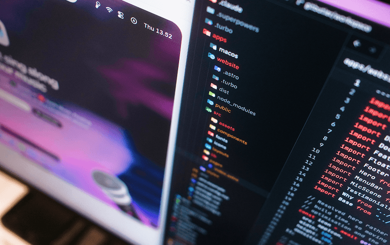 Angled close-up of a developer's dual monitors showing a website preview and a code editor with file structure and import statements.