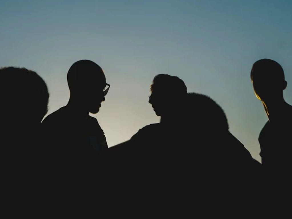 Silhouettes of four people standing together against a twilight sky, suggesting a contemplative or intimate atmosphere, with a soft gradient in the background.