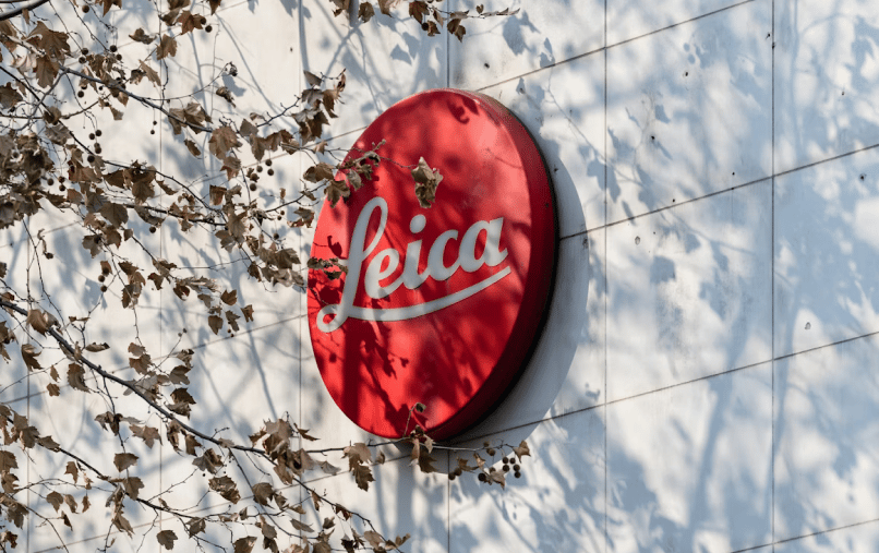 A red Leica logo is mounted on a white tiled wall, partially obscured by the shadows of bare tree branches, conveying a calm, autumnal ambiance.